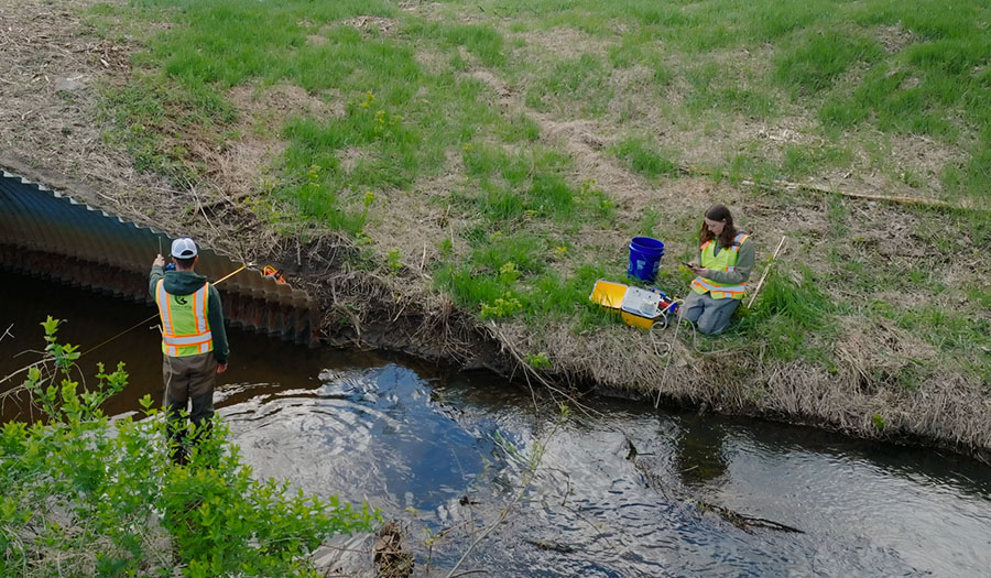 Two watershed staff members collect water quality data from creek. One staff member is standing in the creek while the other staff member kneels on the shoreline.