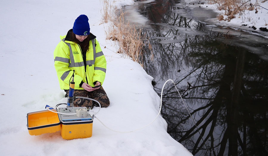 Watershed staff member kneels in the snow next to a creek collecting data on winter chloride levels