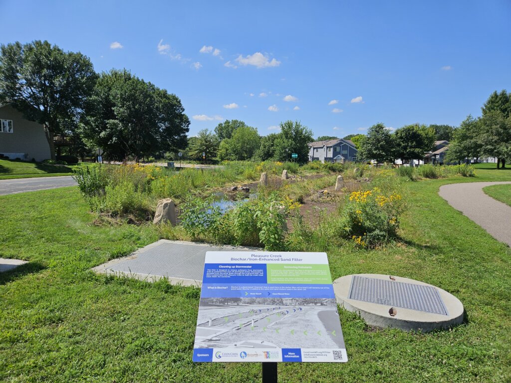 Informational sign in a park describing a biochar/iron‑enhanced sand filter project, with greenery and houses in the background.