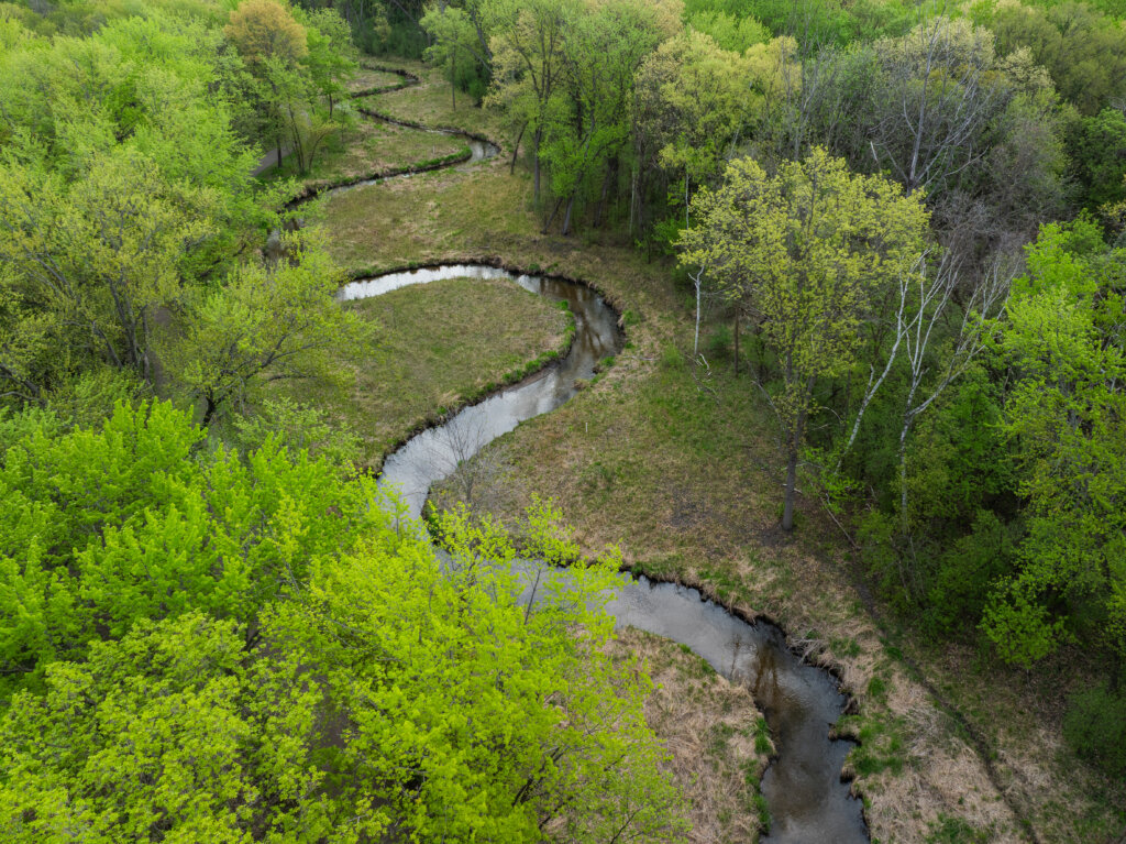 Aerial photo of Sand Creek post restoration. The creek curves through the landscape with trees lining both sides. The landscape suggests the photo was taken in early spring.