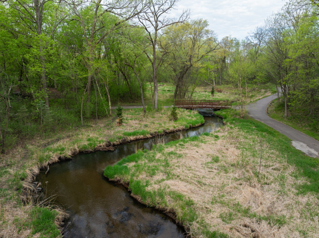 Photo of Sand Creek flowing under a footbridge, post-restoration. A paved walking trail runs along the right side of the creek. The landscape suggests the photo was taken in early spring.
